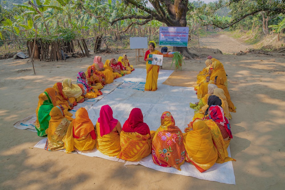A group of women sitting in a semi-circle and a community trainer standing at one end conducting training with the help of a booklet.