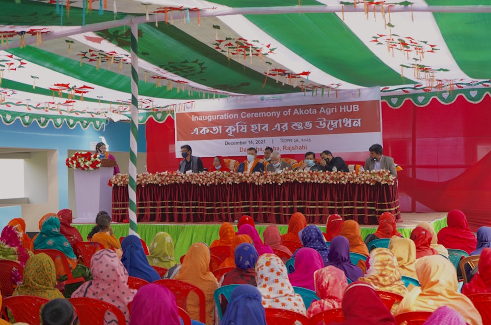 A group of women sit facing a stage, behind which a banner reads