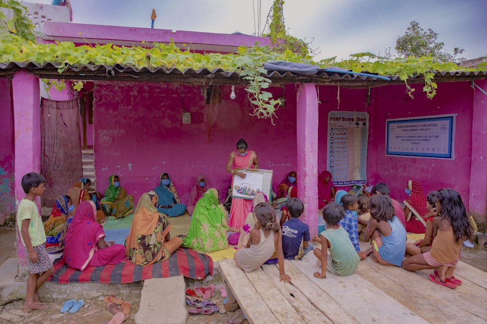 A women holding a learning guide and conducting a training session with a group of women in a village. 