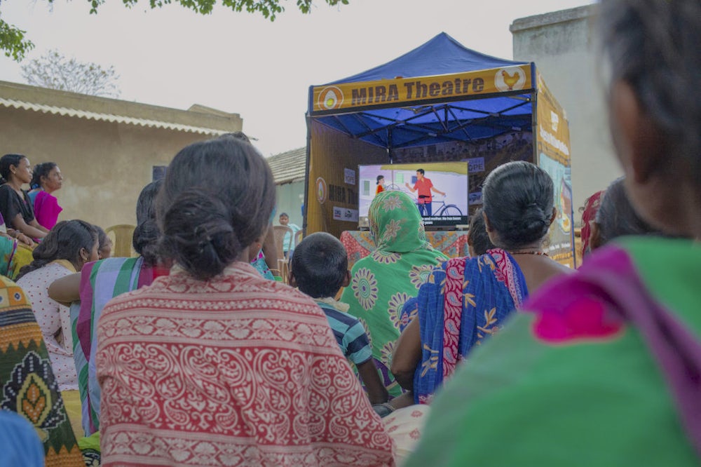A group of men and women sitting and watching an informative movie on a screen in a village.