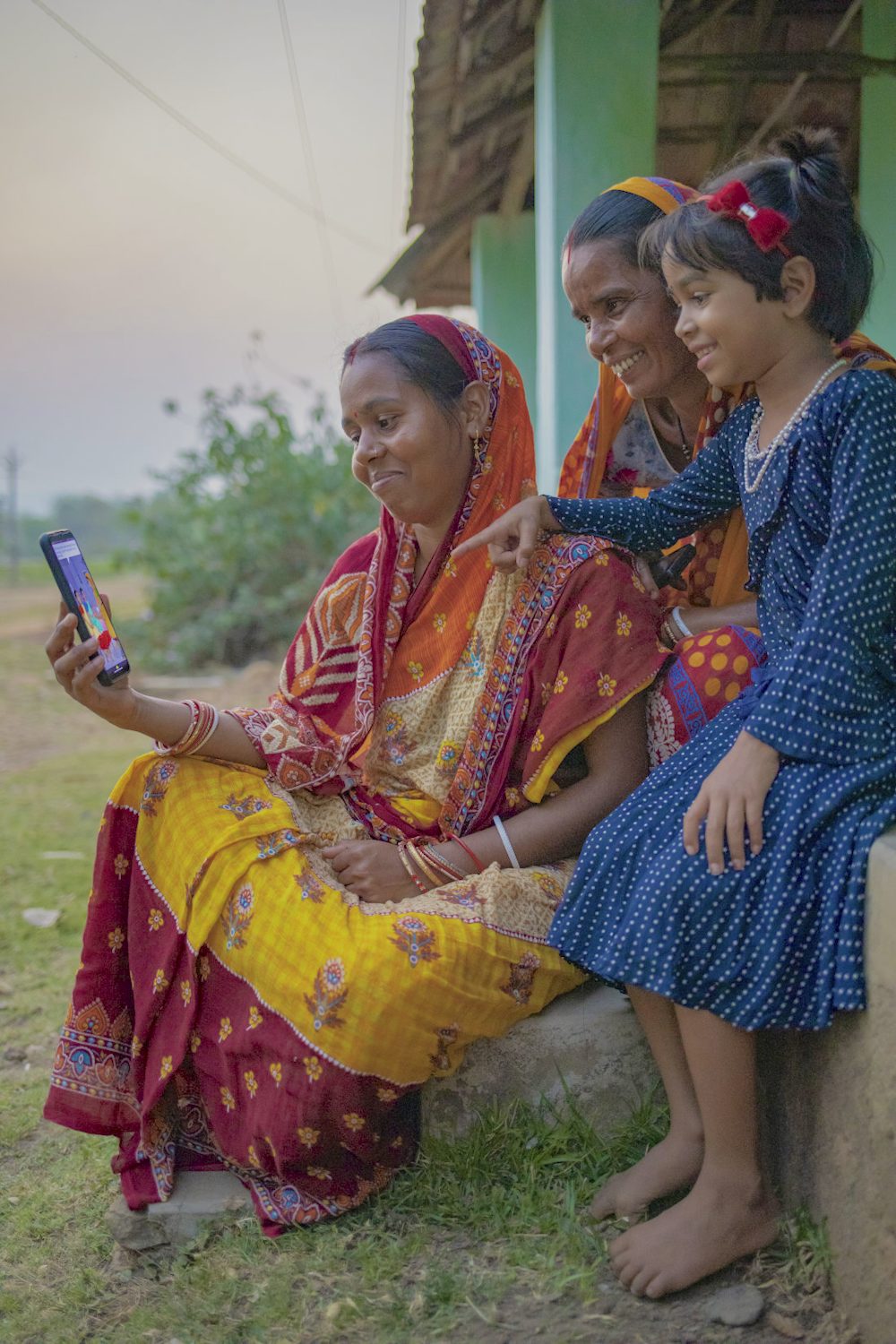 A woman, with her daughter, sitting outside her house in a village and scrolling through a digital lesson on her phone.