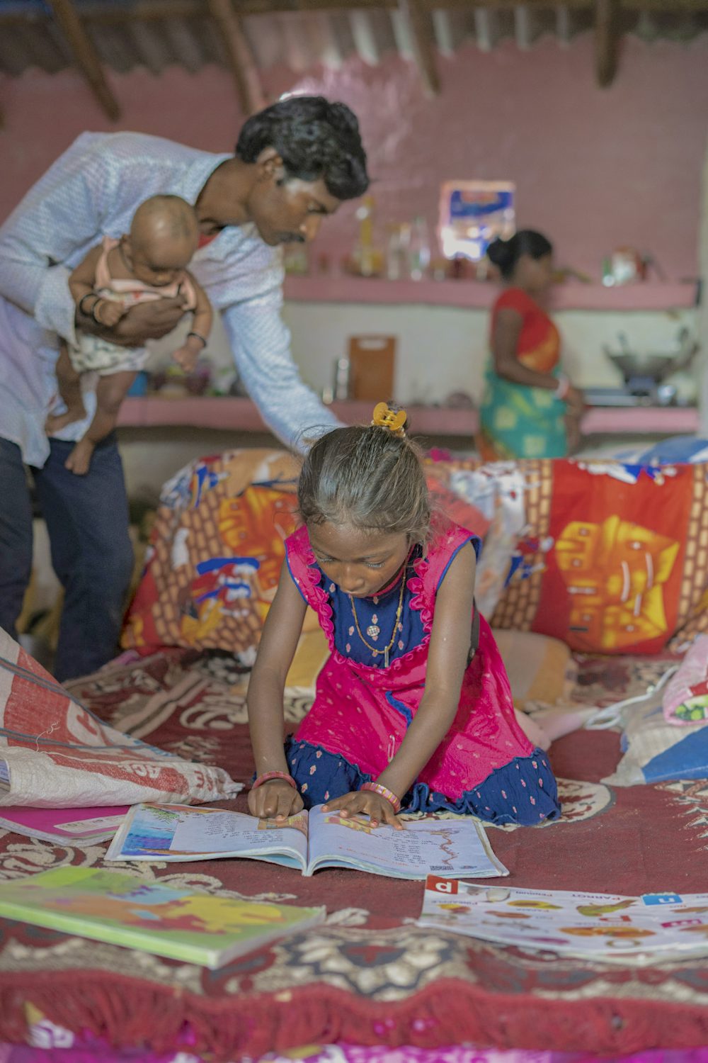 A young girl studying from her notebook while her parents attend household chores in the background. 