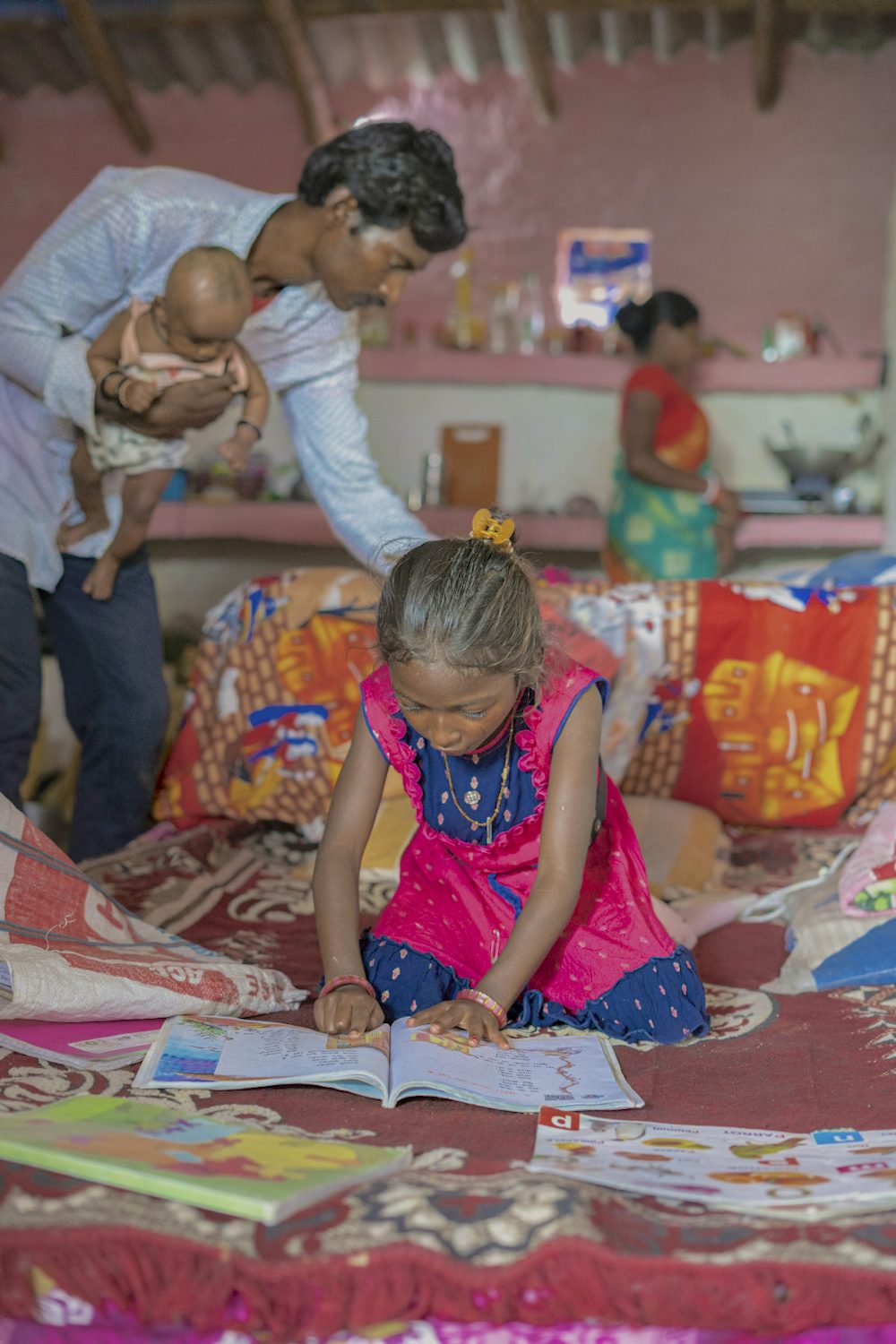 A young girl studying from her notebook while her parents attend household chores in the background.