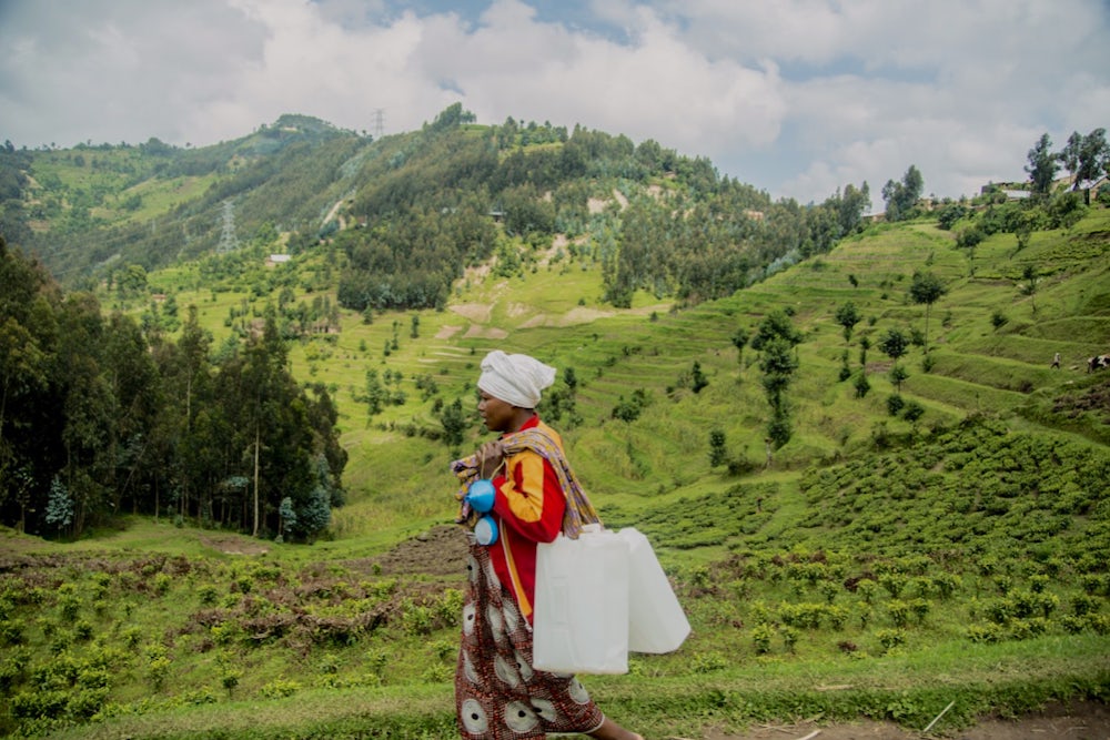 A woman walks through a green hillside with containers over her shoulder.