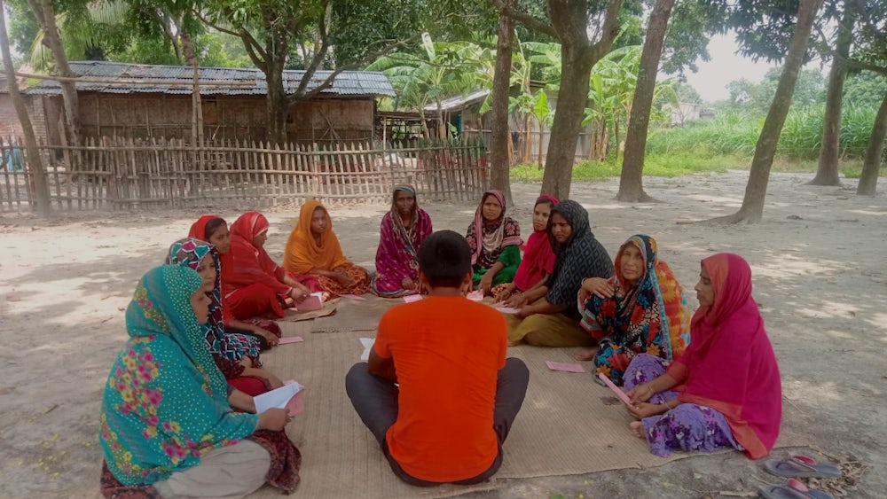 A group of women sitting on the ground in a crescent shape with a man sitting in the middle and delivering a training session.