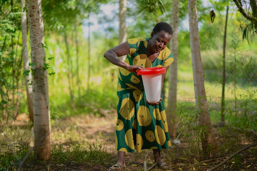 A woman standing in a green field adjusting a water drinker for birds before placing it on the ground. 