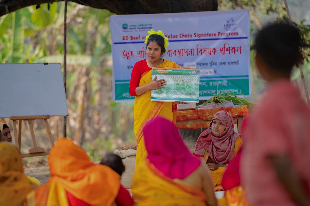 A woman in a vibrant yellow saree conducts a training session outdoors, holding up a chart, with women in colorful attire listening attentively.