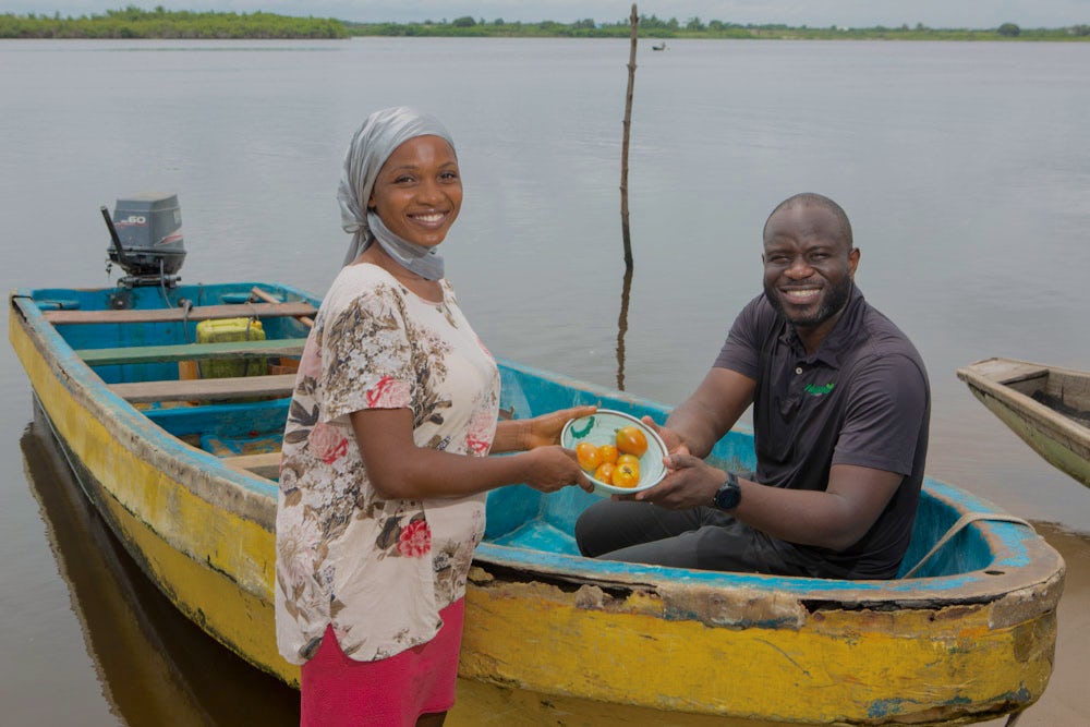 A woman exchanges a bowl of tomatoes with a man inside a boat against a serene river backdrop.