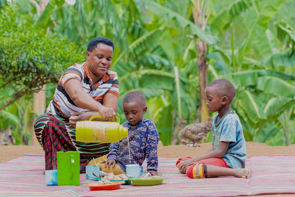 A woman serves milk to her two young children outdoors, surrounded by banana plants.