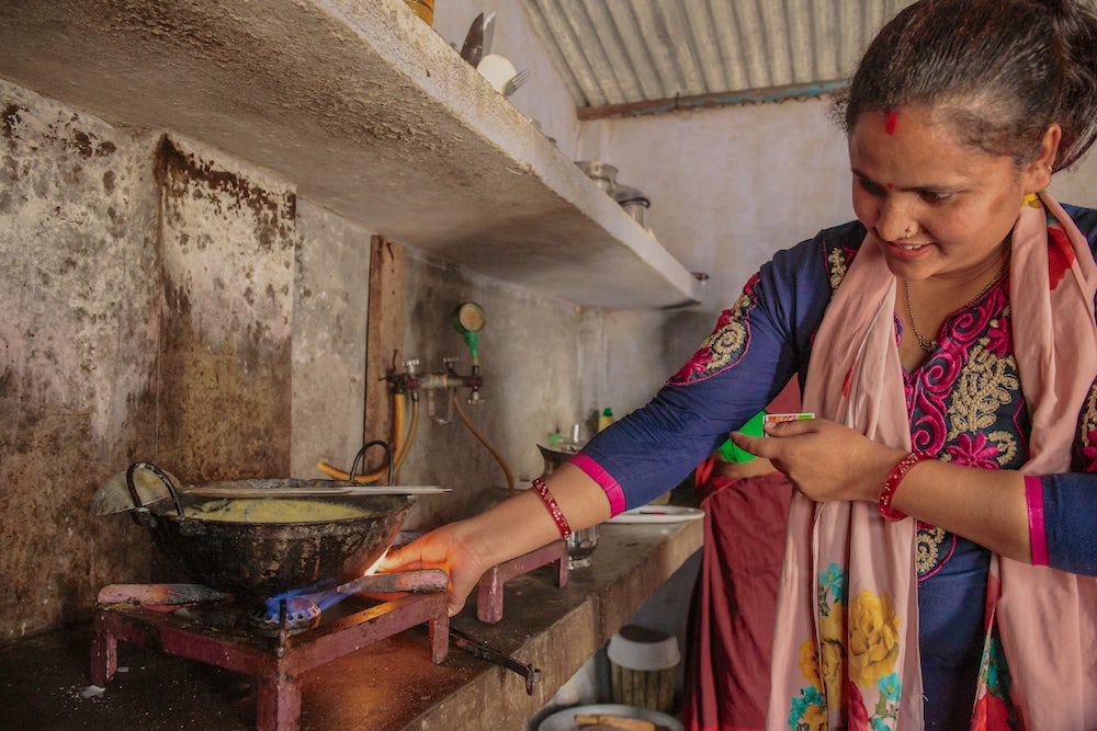 A smiling Indian woman lights her gas stove in her kitchen.