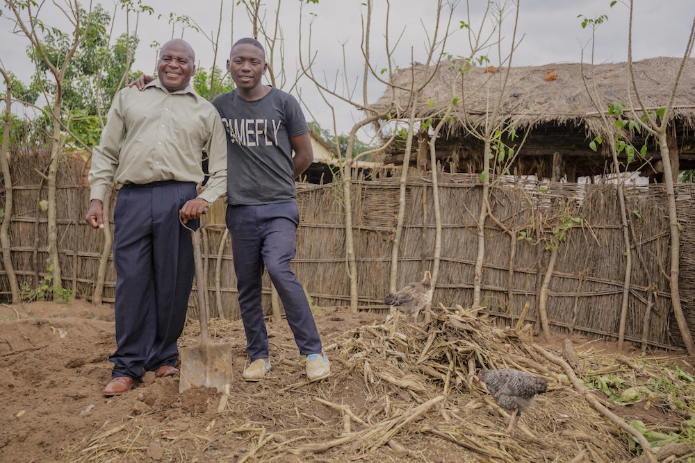A father and son stand side-by-side on their compost pit in Malawi. Both are smiling and the dad holds a shovel in his hand.