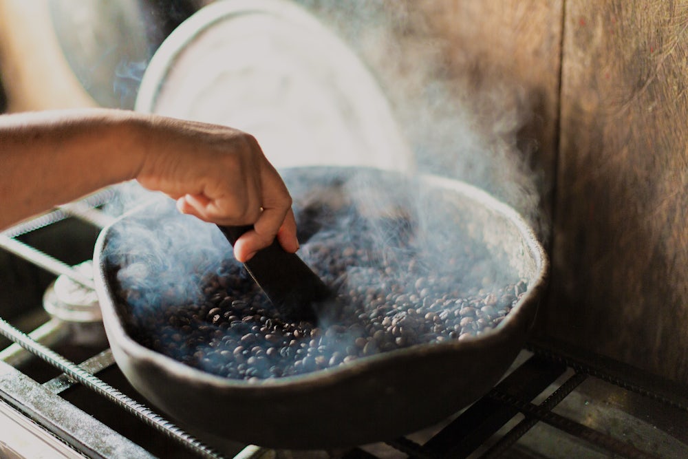 A close up shot of coffee beans roasting in a pan. A pair of hands uses a tool to mix them.
