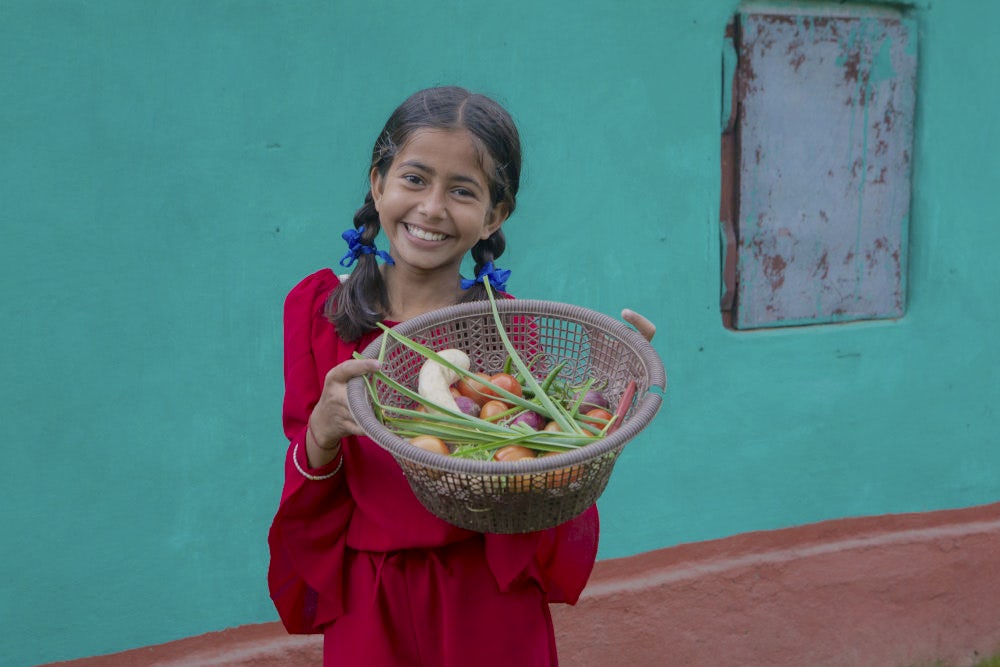 A young girl smiling and holding a basket full of vegetables. 