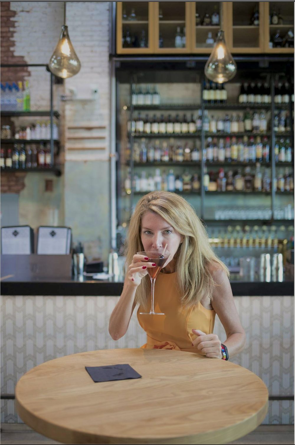 Laura Reiley sits at a bar table sipping a cocktail. She is wearing a yellow dress and looking directly into the camera over the rim of her glass.