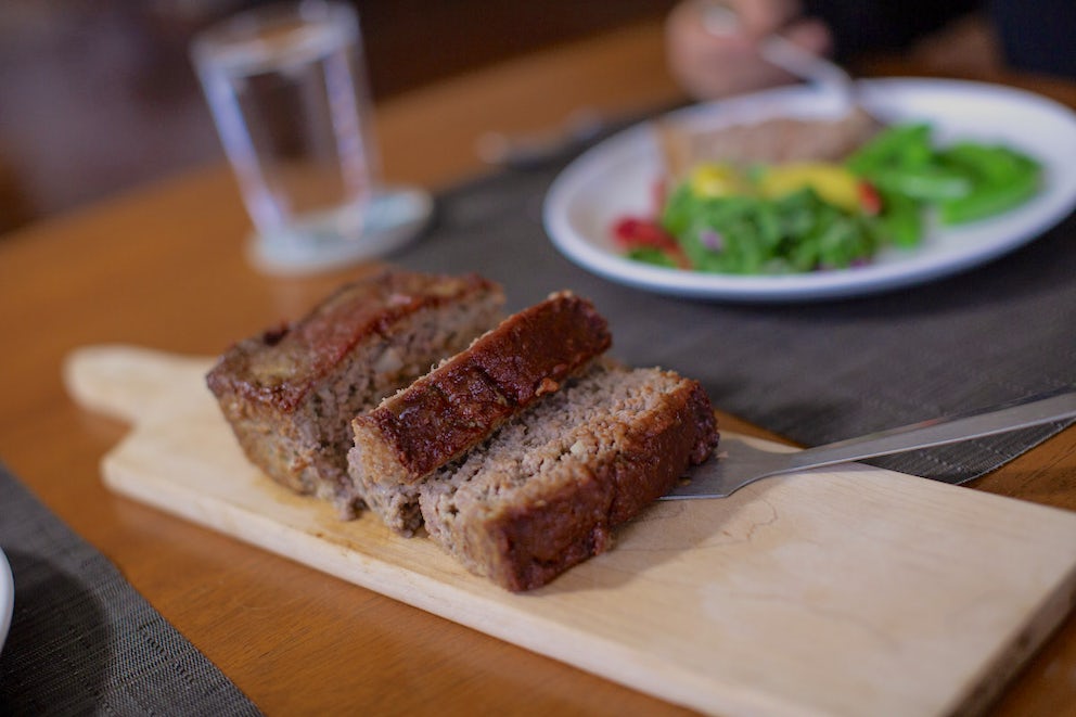 Sliced meatloaf sits on a cutting board. Behind it is a plate filled with green vegetables, waiting for a slice.