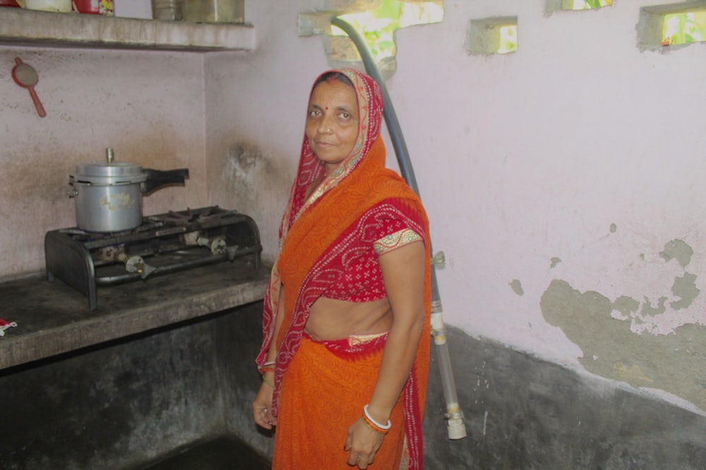 An Indian woman, wearing a bright orange sari, stands next to her stove.