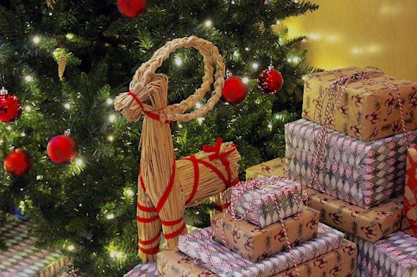 A small goat made of straw sits on top of a pile of wrapped christmas gifts, next to a tree. The goat is decorated with red ribbon.  