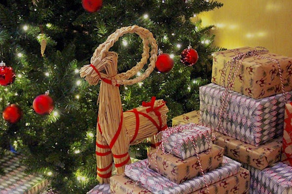 A small goat made of straw sits on top of a pile of wrapped christmas gifts, next to a tree. The goat is decorated with red ribbon.