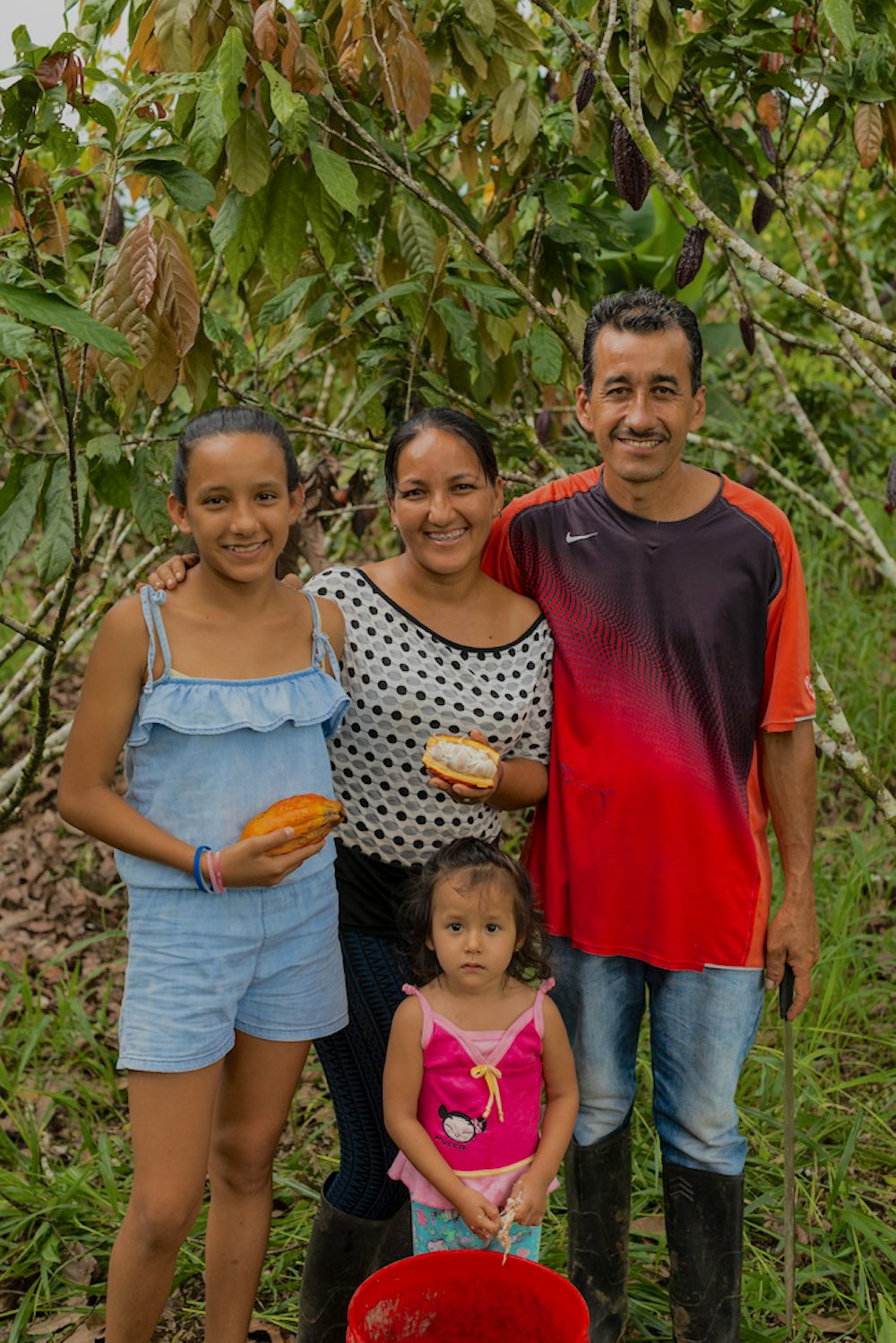 A family portrait in the chocolate grove of a man with his arm around his wife and older daughter, who are holding cacao pods, with the youngest daughter standing in front.