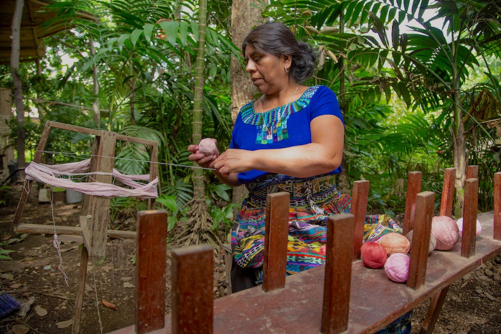 Rosalinda Tay sits outside her studio and winds yarn died with cochineal on her enrollado. In front of her is an urdidora or warping frame.