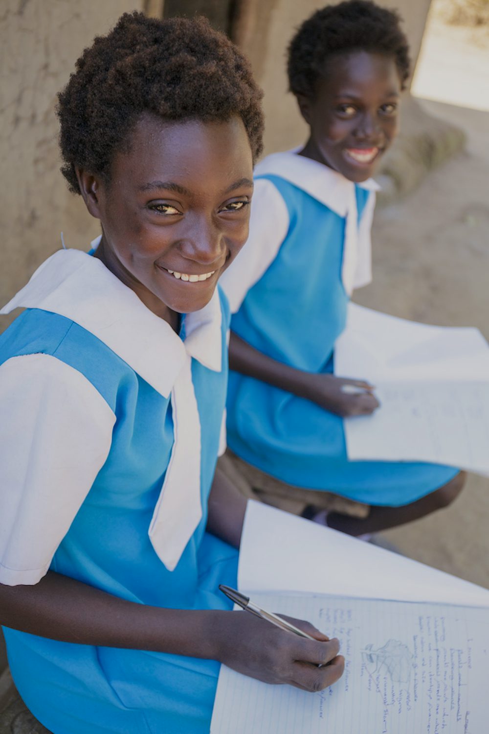A 13 year old girl in a blue and white school uniform looks smiling while writing with a pencil on paper.