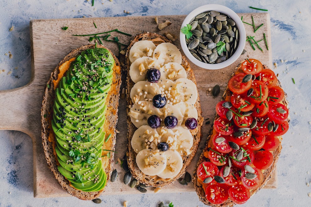 Avocado toast, banana and blueberry toast, and tomatoes on toast on a cutting board.