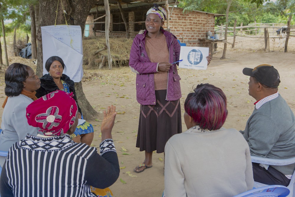 An african woman gives a presentation to a group of men and women outside of her home.