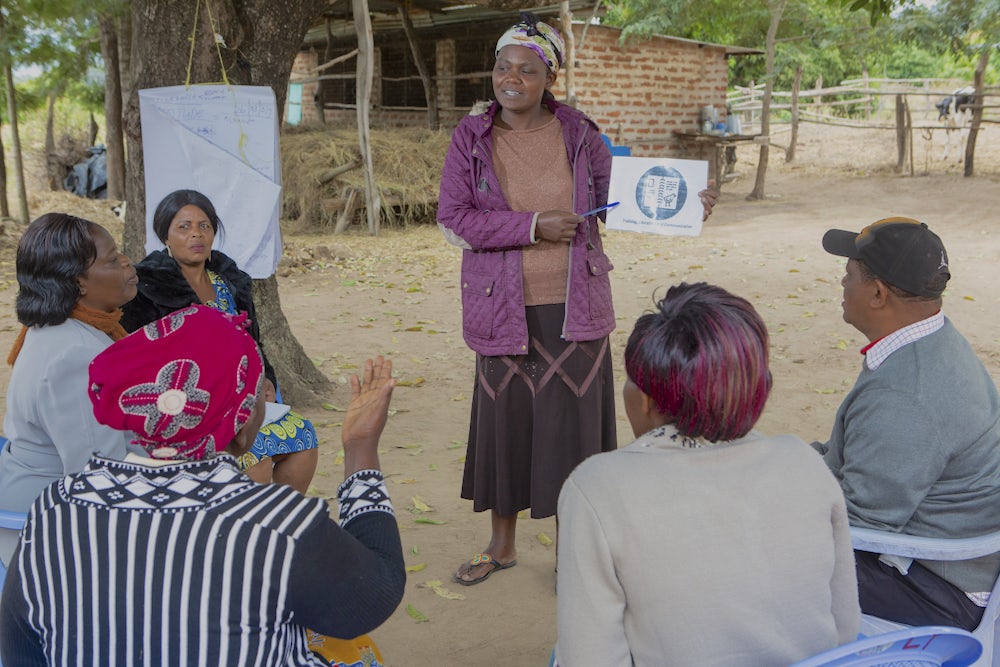 An african woman gives a presentation to a group of men and women outside of her home. 