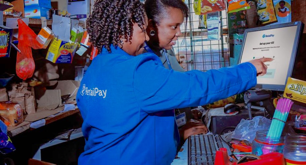 Two women (one wearing a branded retail pay shirt) stand over a computer screen. They are smiling and pointing. 
