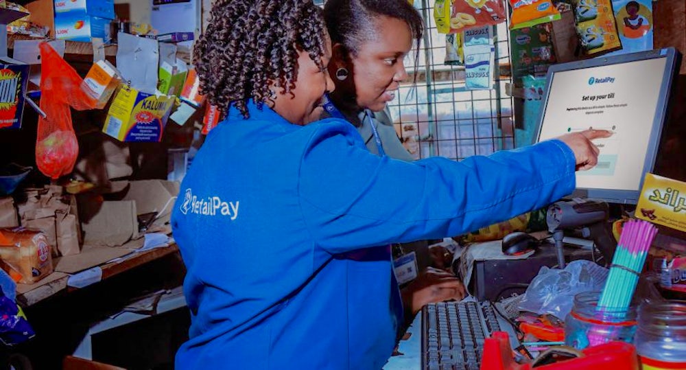 Two women (one wearing a branded retail pay shirt) stand over a computer screen. They are smiling and pointing.