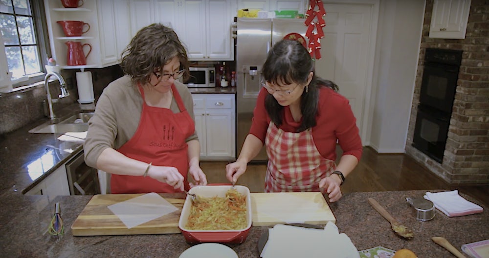 Two women in red aprons look over a pan of spring roll filling.