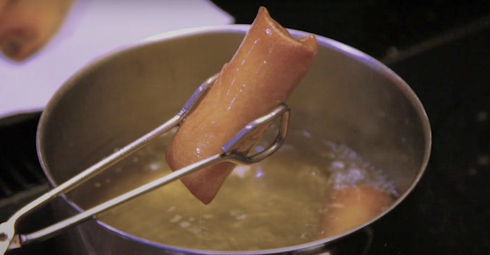 A pair of tongs remove a golden brown spring roll from hot oil. 