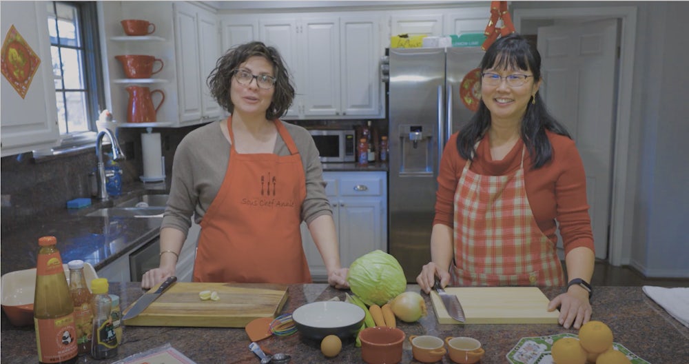 Two women, in red aprons, stand smiling for the camera. They are standing behind a kitchen island.