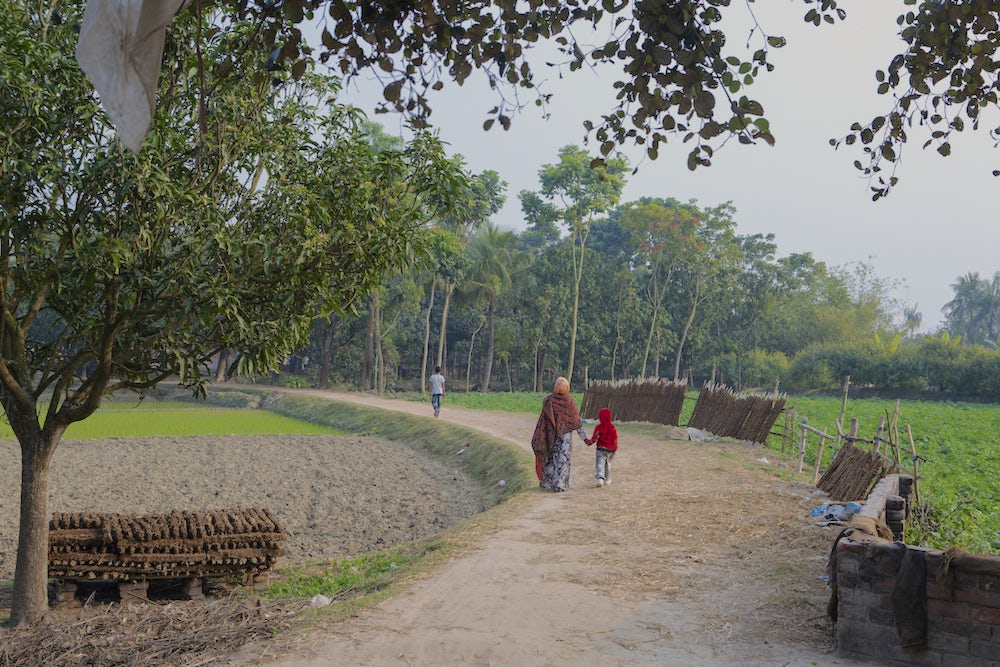 A woman and a small child walk down a dirt road away from the camera. The woman is wearing a bright red sari.