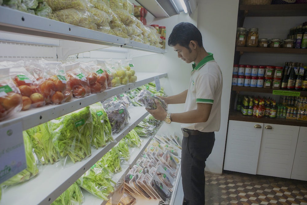A Cambodian man stands in front of a refrigerated produce section of a local grocery store holding a fresh vegetable.
