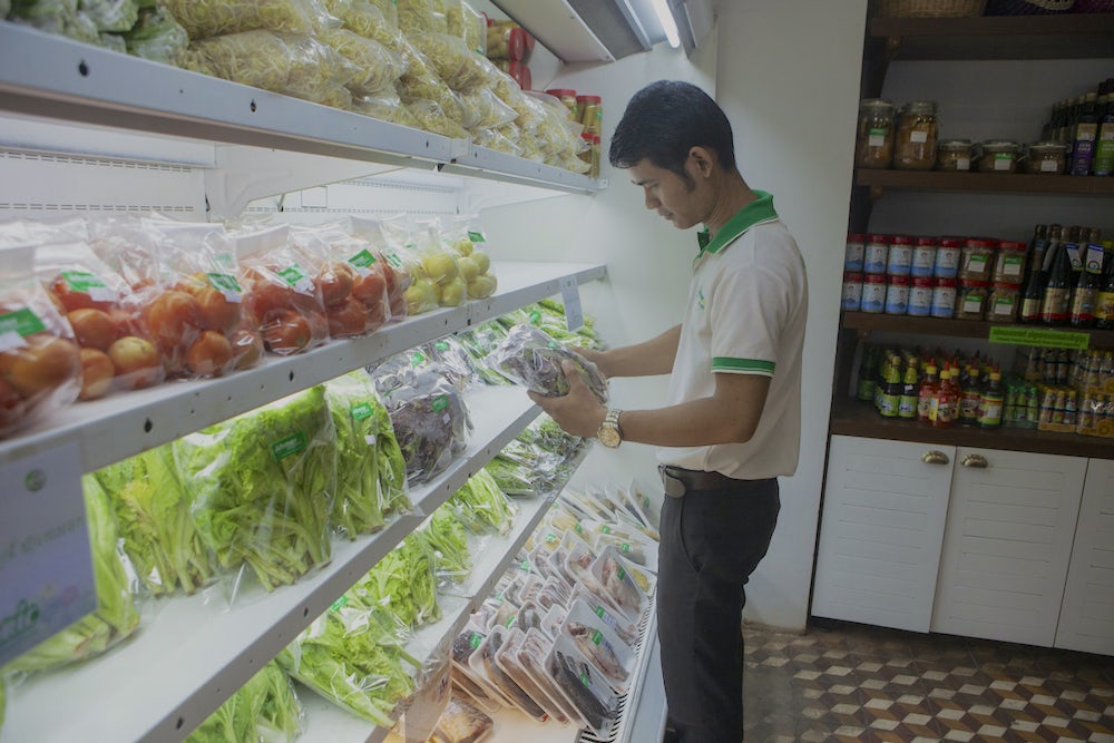 A Cambodian man stands in front of a refrigerated produce section of a local grocery store holding a fresh vegetable. 