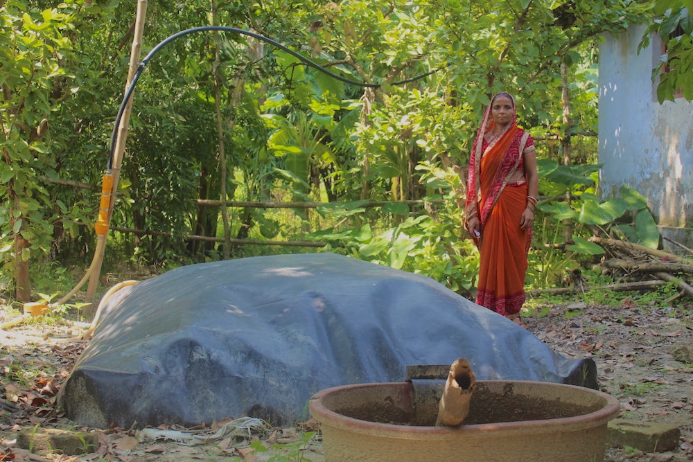An Indian woman in orange dress stands behind her biodigester, a large grey dome on the ground.