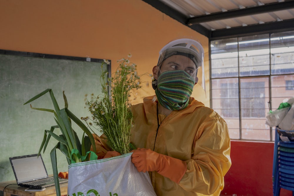 A man wearing a protective face shield and gloves carries a bag of produce.