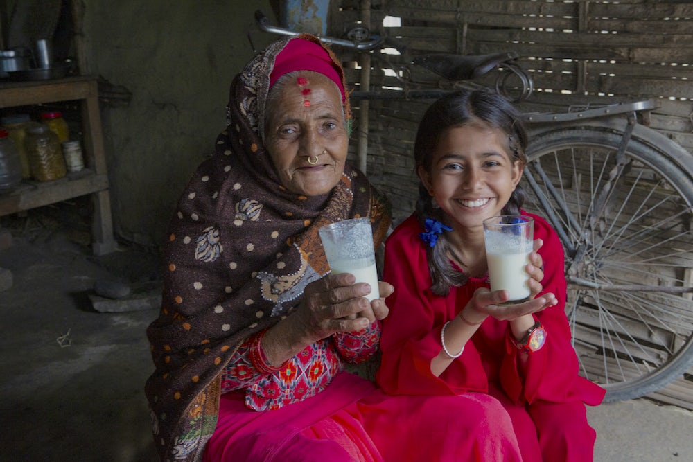 A smiling Nepali woman in her 70s and her 12-year-old granddaughter each hold a glass of milk in their hands.