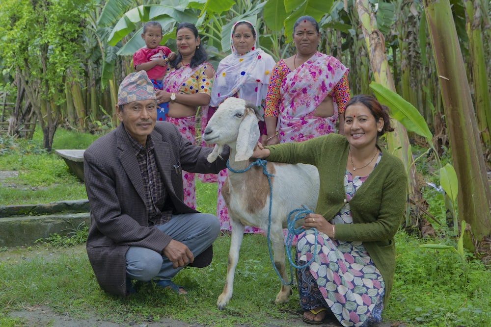 A woman and man crouch next to their goat. Four women stand behind them, smiling.