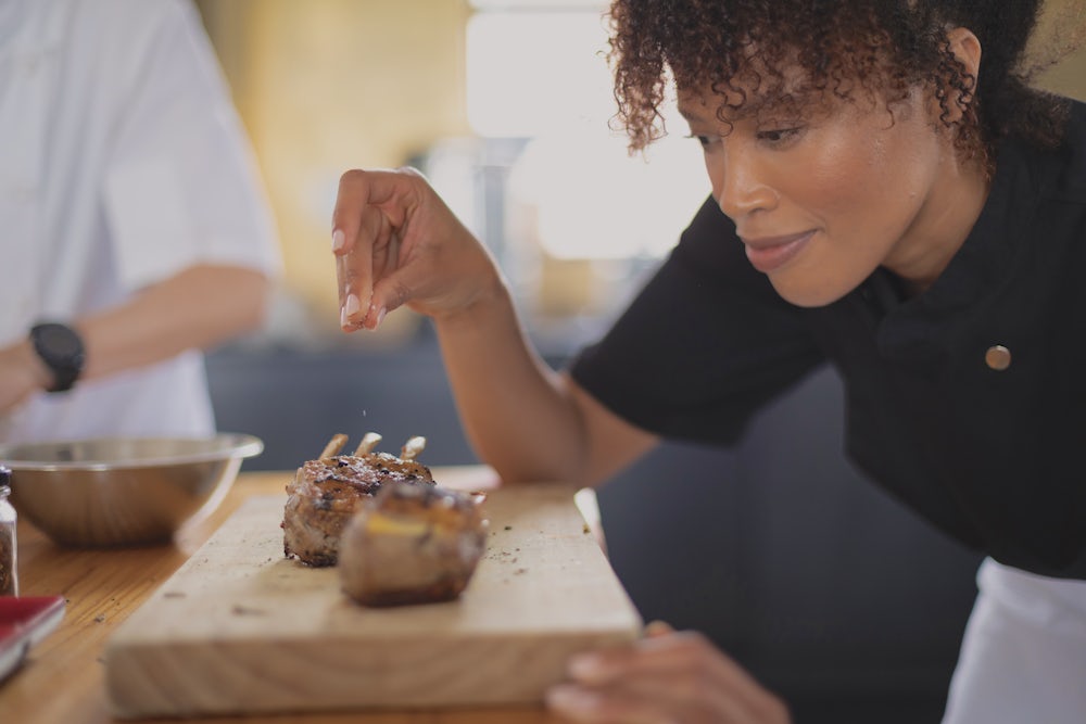 A woman chef seasons two lamb chops.