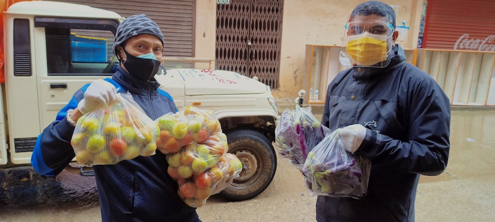 Two men wearing masks and face shields hold bags of produce.