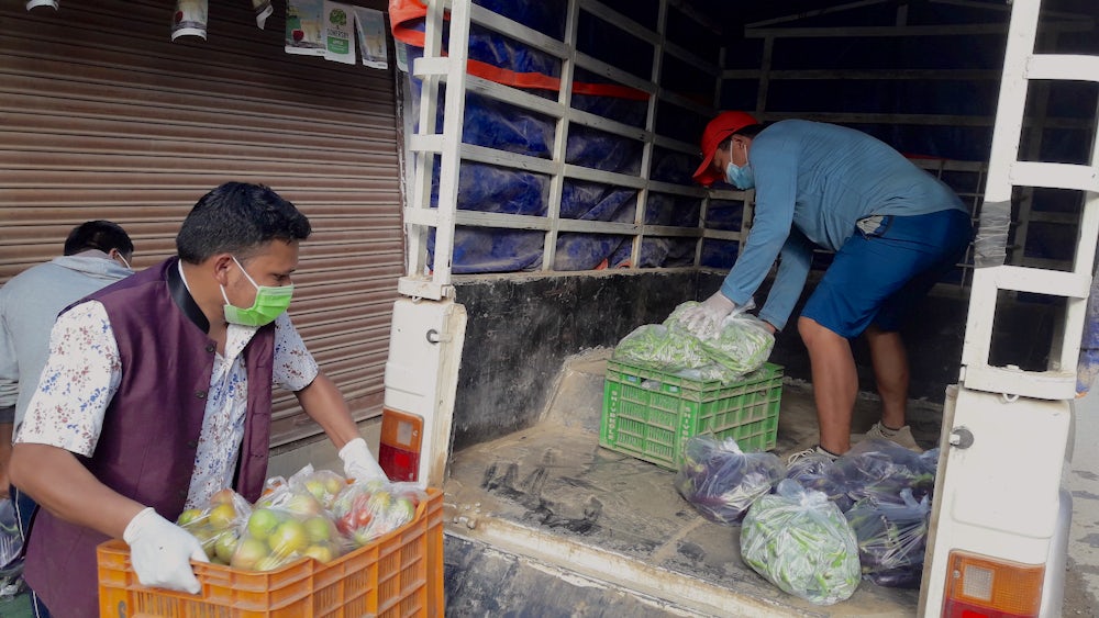 Two men wearing personal protective equipment load fresh vegetables into the back of a truck.