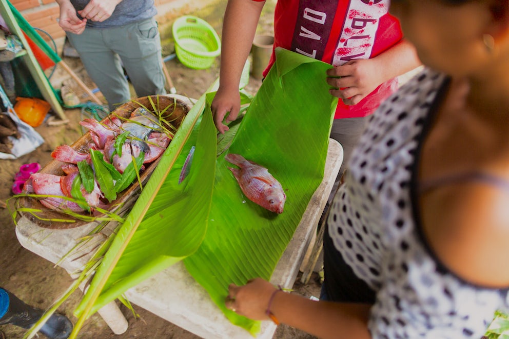 Top-down view of a woman wrapping a red whole tilapia in a banana leaf, next to a wooden bowl full of fish with cilantro on top.