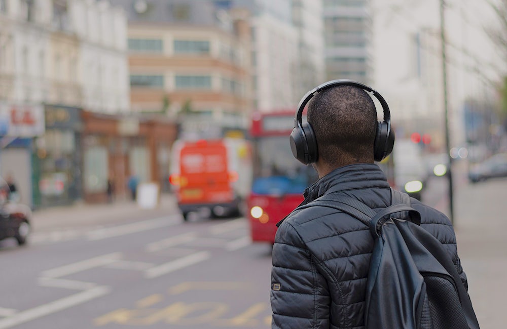 A man stands with his back to the camera near the crosswalk of a busy, city street. There are cars in the background of the picture and he is wearing headphones, a puff jacket and a backpack.