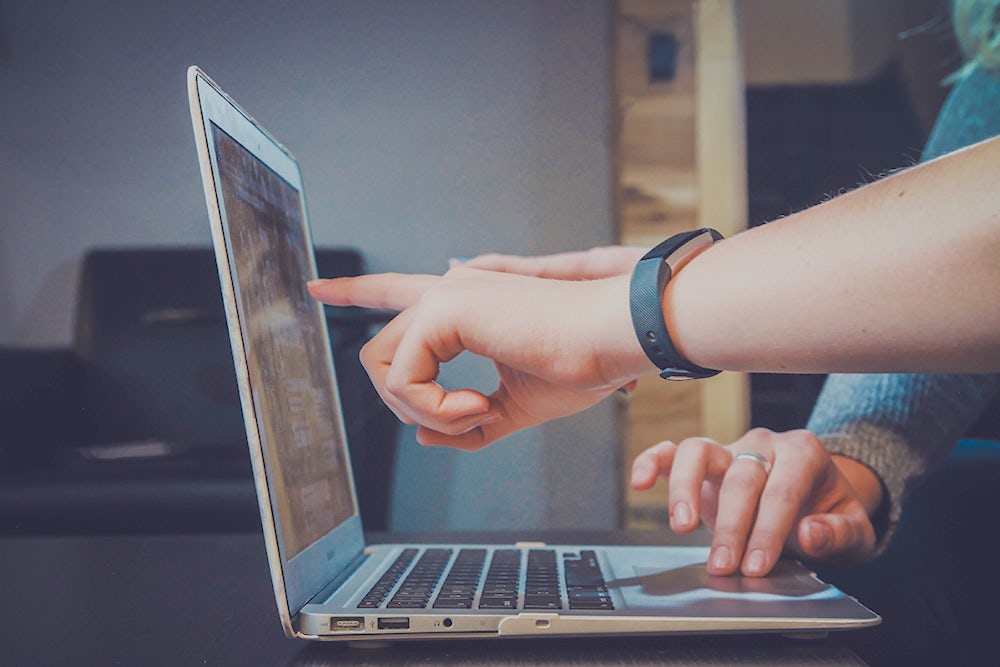 Profile view of a laptop with two pairs of hands. One is pointing at the screen as if it's seen something interesting. The other is using the trackpad.