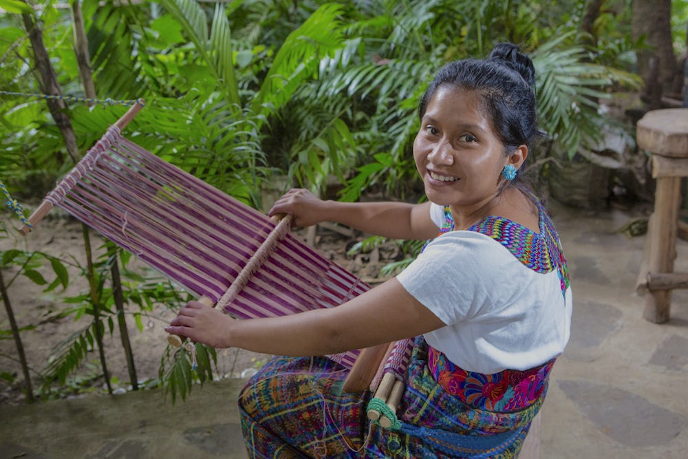 A woman sits at a backstrap loom weaving a shawl with various shades of crimson. She is smiling at the camera.