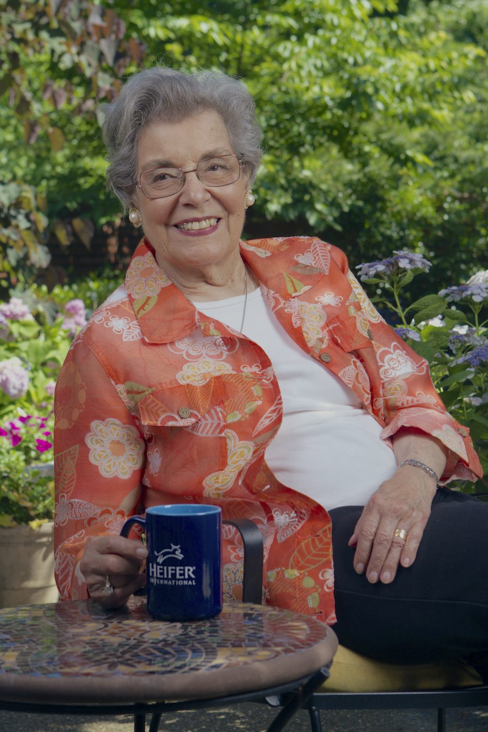 A smiling woman in her 70s sits in a chair holding a Heifer International coffee cup.
