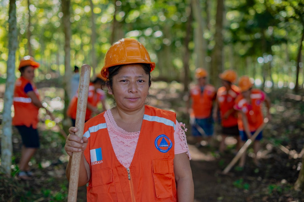 A Guatemalan woman with a bright orange vest stands holding a shovel in a forest, working with a team to reduce forest fire risk.