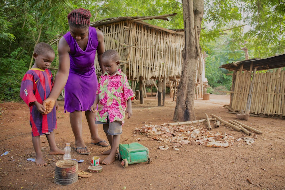 A woman and her two young sons play barefoot on a dirt floor with toys made from plastic jugs and tin cans.